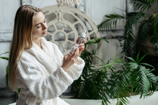 Smiling young woman in white bathrobe looking at bottles with facial cleanser and natural organic cosmetics for skin. Serene female spending morning in bathroom, make daily skincare routine at home - Фото, зображення