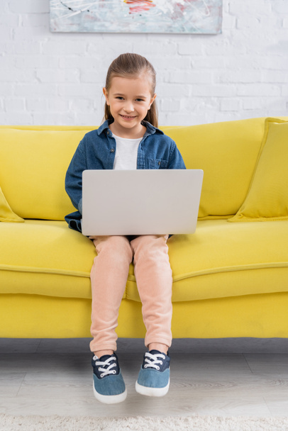 Kid with laptop smiling at camera on yellow sofa  - Φωτογραφία, εικόνα