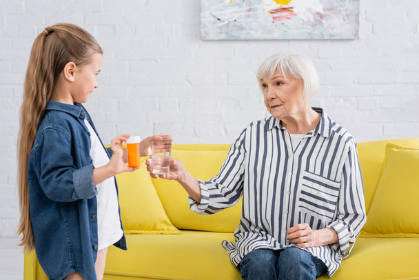 Осторожная девушка держит таблетки и воду рядом с бабушкой на диване  - Фото, изображение