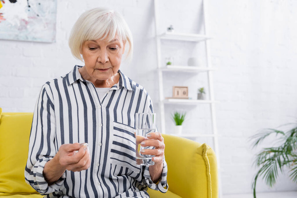 Donna anziana sconvolta che tiene pillola e bicchiere d'acqua in soggiorno  - Foto, immagini