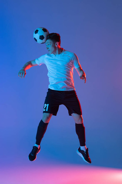 Young Caucasian man, male soccer football player jumping isolated on gradient blue pink background in neon light. Back view - Fotografie, Obrázek