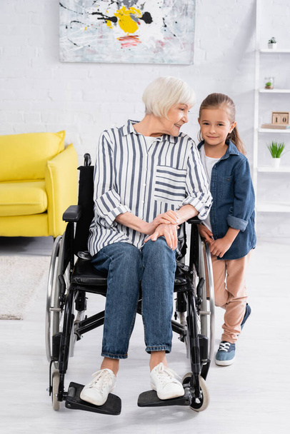 Smiling woman in wheelchair looking at granddaughter at home - Fotó, kép