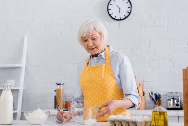 Vanhempi nainen esiliina ottaen jauhot lähellä maitoa ja munia keittiössä  - Valokuva, kuva