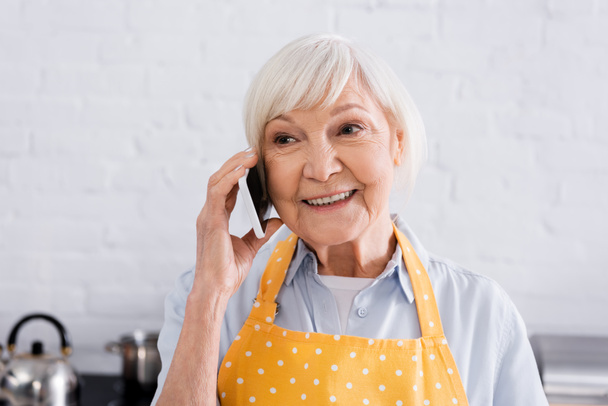 Mujer anciana sonriente en delantal hablando en smartphone en la cocina  - Foto, Imagen