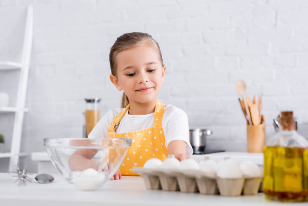 Kid in apron standing near blurred eggs and oil in kitchen  - Fotografie, Obrázek