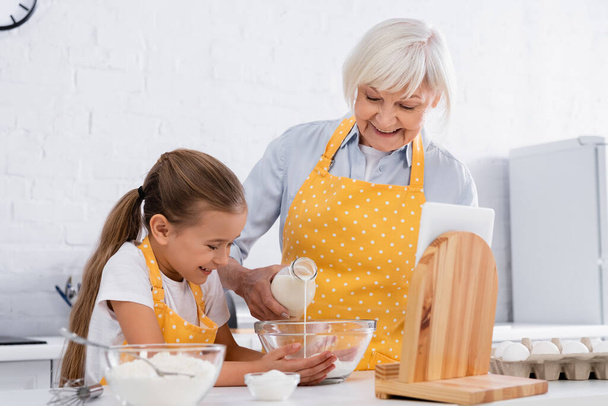 Positive kid and grandmother pouring milk in bowl near digital tablet and flour  - Photo, Image
