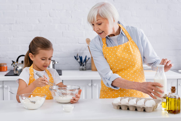 Smiling granny holding bottle of milk near kid, eggs and flour in kitchen  - Photo, Image