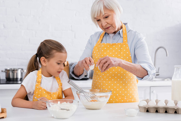 Mujer mayor vertiendo huevo en tazón cerca de los ingredientes y nieta en la cocina  - Foto, imagen