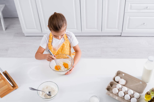 Vista aerea di bambino in grembiule che mescola uova in ciotola in cucina  - Foto, immagini