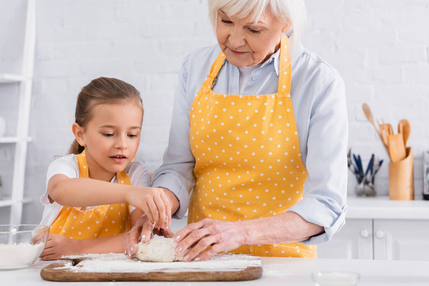 Kid and granny in aprons making dough together in kitchen  - Фото, изображение