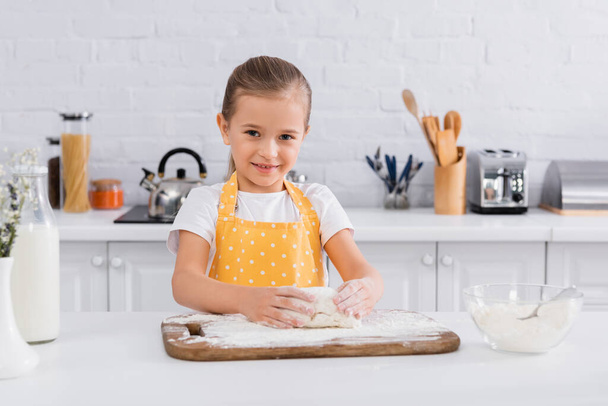 Happy girl in apron preparing dough near ingredients  - Фото, изображение