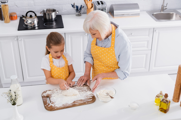 Overhead view of smiling granny helping to child with dough in kitchen  - Foto, Bild