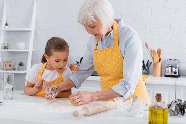 Granny and granddaughter standing near dough and cookie cutter in kitchen  - Φωτογραφία, εικόνα