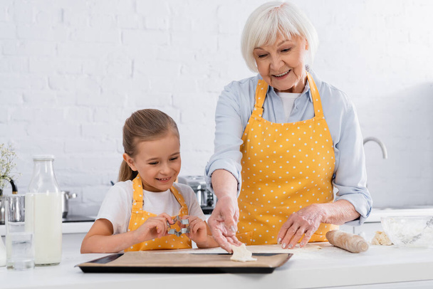 Smiling kid holding cookie cutter near granny with dough  - Foto, Bild