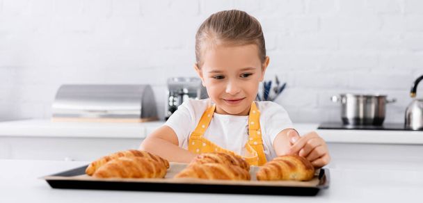 Hymyilevä tyttö esiliinassa koskettaa paistettua croissantia keittiössä, banneri  - Valokuva, kuva