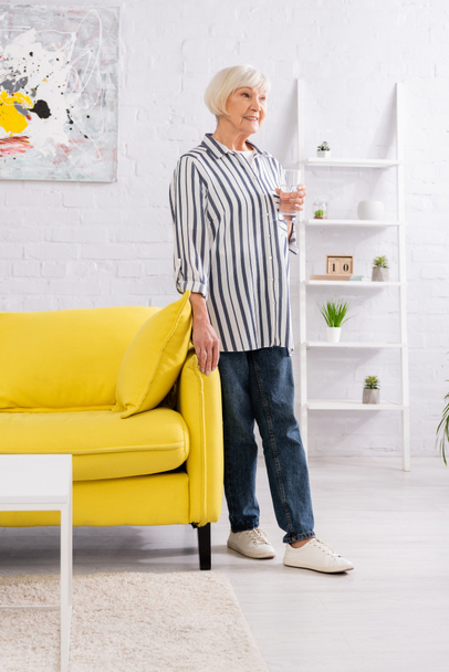 Happy elderly woman with glass of water standing in living room  - Foto, imagen