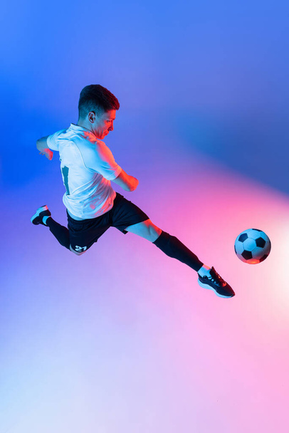 Genç bir adamın yüksek açılı görüntüsü, erkek futbolcu eğitimi neon ışıklarıyla izole edilmiş mavi bir arka planda.. - Fotoğraf, Görsel