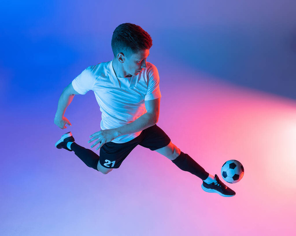 High angle view of young man, male soccer football player training isolated on gradient blue pink background in neon light. - Fotó, kép