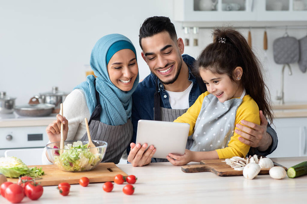 Felice famiglia musulmana utilizzando tavoletta digitale durante la cottura in cucina insieme - Foto, immagini