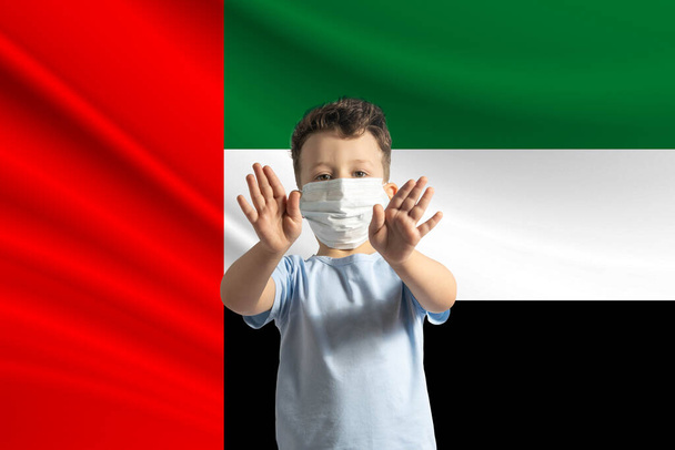 Μικρό λευκό αγόρι με προστατευτική μάσκα στο φόντο της σημαίας των Ηνωμένων Αραβικών Εμιράτων. Κάνει στοπ με τα χέρια του, μένει στο σπίτι Ηνωμένα Αραβικά Εμιράτα. - Φωτογραφία, εικόνα