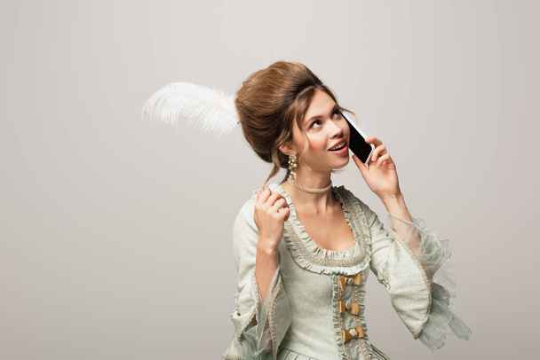 femme rêveuse et souriante en robe rétro touchant les cheveux tout en parlant sur téléphone portable isolé sur gris - Photo, image