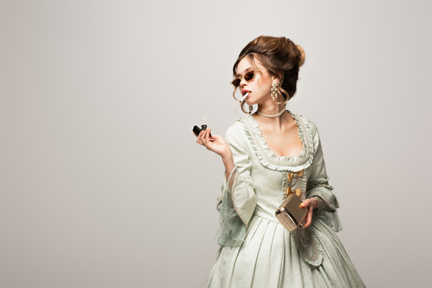 mujer sensual en elegante vestido vintage sosteniendo bolso de embrague dorado mientras fuma aislado en negro - Foto, imagen