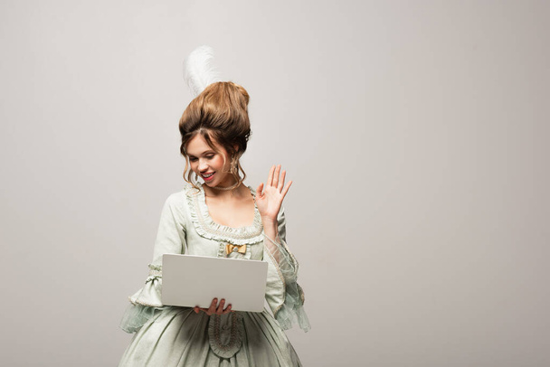 усміхнена жінка в ретро-сукні махає рукою під час відеодзвінка на ноутбук ізольовано на сірому
 - Фото, зображення