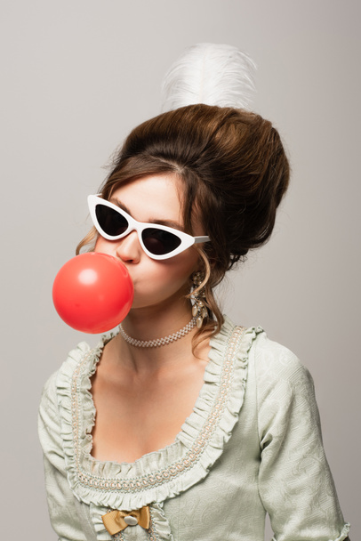винтажный стиль женщины в модных солнцезащитных очках, дующий красный пузырь жвачки изолированы на серый - Фото, изображение