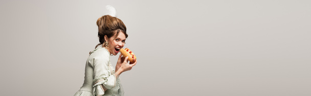 hungrige, im Retro-Stil gehaltene Frau, die in die Kamera schaut, während sie vereinzelt Hot Dog auf grauem Grund isst, Banner - Foto, Bild