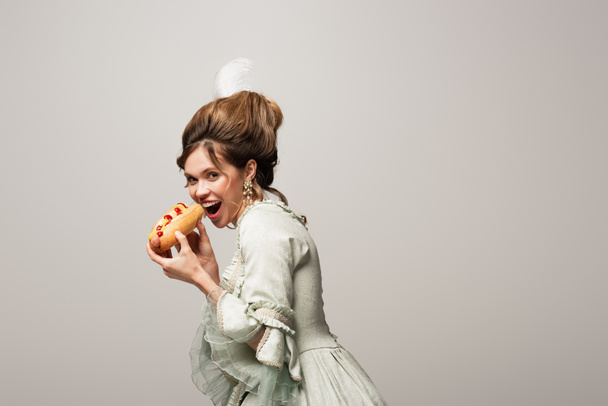 hungry woman in vintage outfit eating tasty hot dog isolated on grey - Photo, Image