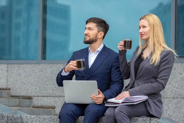 successful businessmen man and woman sit on the stairs of a business building with documents and a laptop in their hands, drink coffee and look at the beauty of the cit - Photo, Image