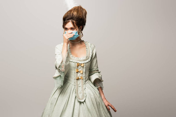 young woman in elegant vintage dress adjusting medical mask isolated on grey - 写真・画像