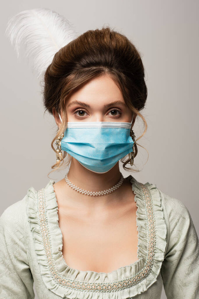 молода елегантна жінка в старовинному вбранні та медичній масці дивиться на камеру ізольовано на сірому
 - Фото, зображення