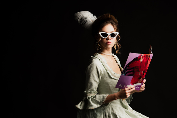 стильная женщина в ретро-платье и модные солнцезащитные очки держа современный журнал изолирован на черном - Фото, изображение