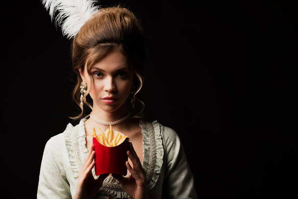 красивая женщина в стиле ретро смотрит в камеру, держа картошку фри изолированной на черном - Фото, изображение