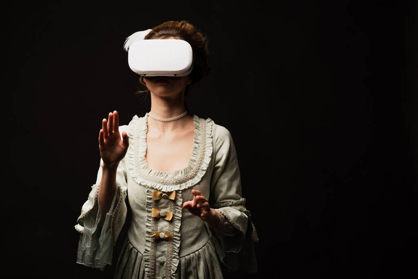stylish woman in elegant vintage dress gaming in vr headset isolated on black - Photo, image
