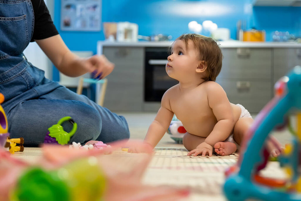 Niedliche entzückende kleine lustige Säugling kaukasischen Neugeborenen Baby sitzt auf dem Boden und genießen Spielzeug und Rasseln mit Mutter in Küche oder Wohnzimmer zu Hause. Kindertagespflege und gesundes Entwicklungskonzept - Foto, Bild