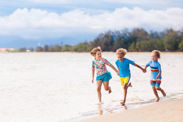 Tropik plajda oynayan çocuklar. Çocuklar yaz tatilinde yüzer ve denizde oynarlar. Kum ve su eğlencesi, küçük çocuklar için güneş koruması. Küçük çocuk ve kız okyanus kıyısında koşup zıplıyorlar.. - Fotoğraf, Görsel