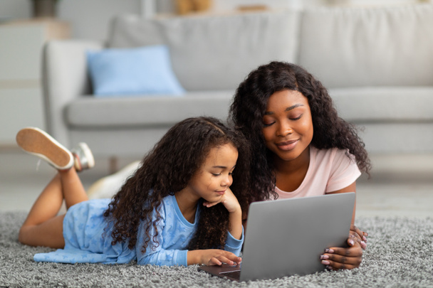 遠隔教育。陽気な黒お母さん助けて彼女の小さな娘ともにウェブレッスンのラップトップコンピュータ上のホーム - 写真・画像