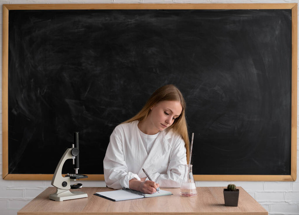 Μια δασκάλα με λευκό παλτό κάθεται στο φόντο ενός καθαρού πίνακα κιμωλίας. Το μικροσκόπιο είναι στο τραπέζι. Η κυρία γράφει σε ένα σημειωματάριο.. - Φωτογραφία, εικόνα