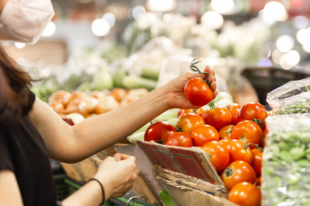 Женщина в защитной маске выбирает томато в овощном магазине. - Фото, изображение