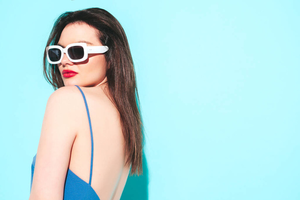 High fashion portrait of young beautiful brunette woman wearing nice summer dress.Sexy trendy model posing near blue wall in studio.Fashionable female looking at camera in sunglasses. Red lips - 写真・画像
