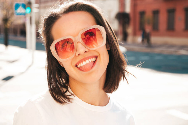 Closeup portrait of young beautiful smiling hipster woman in trendy summer clothes. Sexy carefree woman posing on the street background at sunset. Positive model shows tongue .Happy and cheerful. - Foto, afbeelding