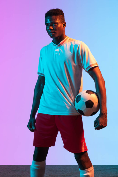 Half-length portrait of African professional football player standing isolated on gradient blue pink background in neon light. - Photo, Image