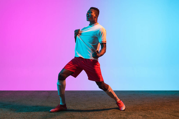 One African man, professional football player standing isolated on gradient blue pink background in neon light. - Фото, изображение