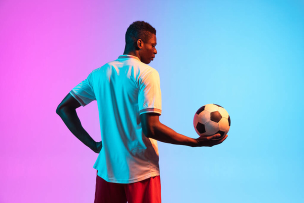 Half-length portrait of African professional football player standing isolated on gradient blue pink background in neon light. Back view - Fotoğraf, Görsel