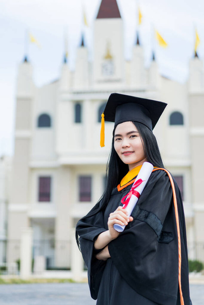 A young beautiful Asian woman university graduate in graduation gown and mortarboard holds a degree certificate stands in front of the university building after participating in college commencement - Photo, Image