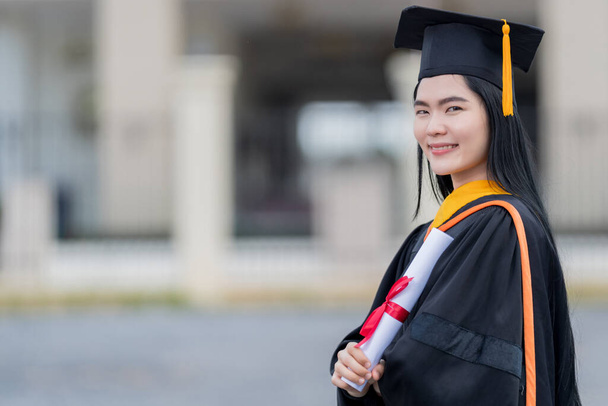 Uma jovem bela mulher asiática universidade graduado em vestido de graduação e argamassa detém um certificado de grau fica em frente ao edifício da universidade depois de participar no início da faculdade - Foto, Imagem