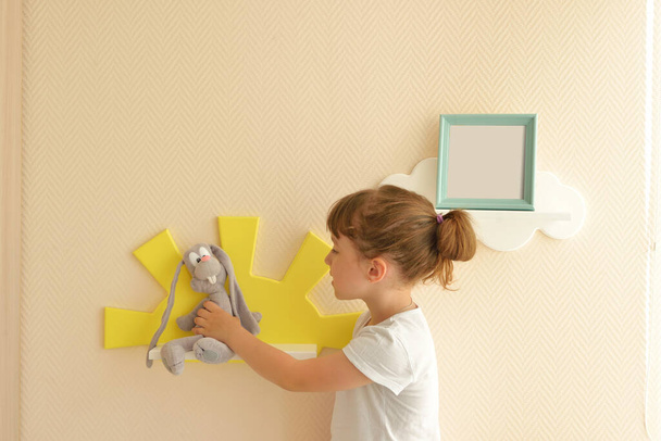 girl happy lifestyle puts a gray hare on a shelf in the form of a yellow sun. Children's shelves in the form of white clouds on a plain beige wall. trendy shelves in the room - Photo, Image