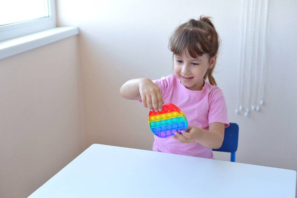 een meisje in een roze t-shirt speelt met een trendy speeltje pop it. kleurrijke antistress zintuiglijke speelgoed fidget duwen pop het in handen van het kind, kan worden gebruikt voor de opleiding met autistische mensen - Foto, afbeelding